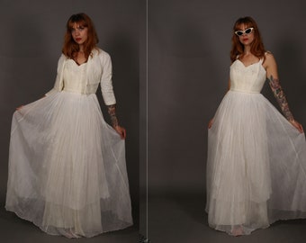May include: A white wedding dress with a fitted bodice and a full, pleated skirt. The dress is layered with tulle and has a lace overlay. The model is wearing a white jacket over the dress.