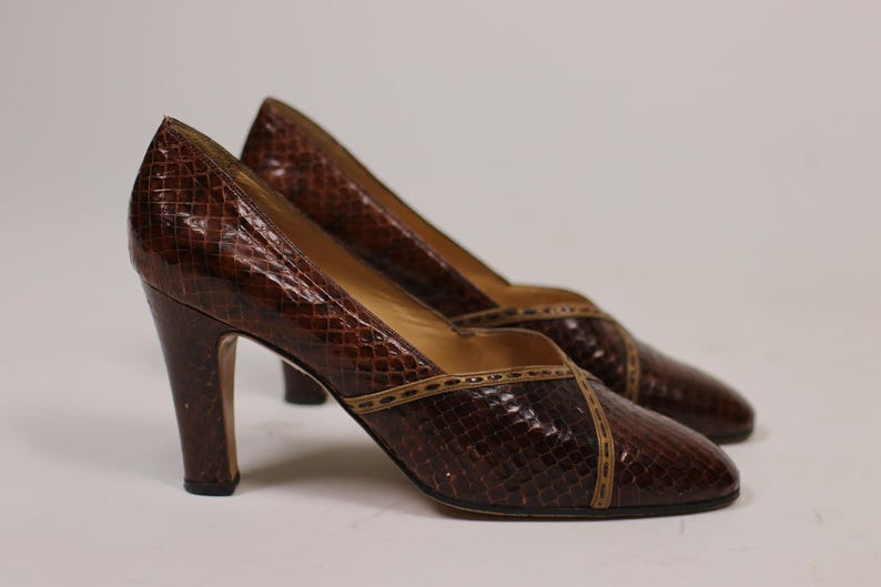 K&ouml;nnte beinhalten: Ein Paar braune Schlangenlederschuhe mit braunem Lederbesatz. Die Schuhe haben eine spitze Zehenpartie und einen hohen Absatz.