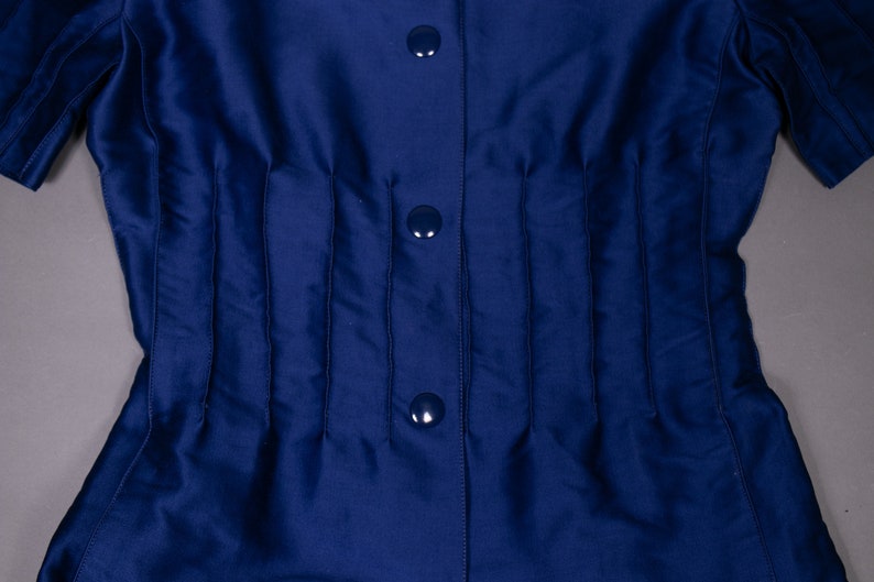 May include: A navy blue shirt with a button-up front and a series of vertical pleats. The shirt has three buttons.