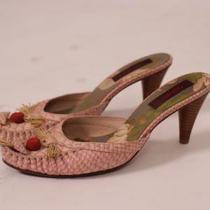 May include: A pair of pink, open-toe mules with a textured pattern. The shoes have a high heel, decorative tassels, and red pom-poms on the front. The interior features a floral design.