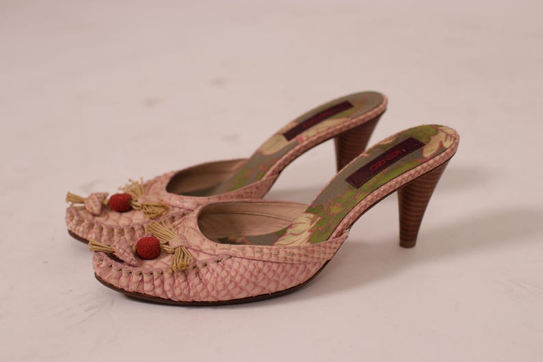 May include: A pair of pink, open-toe mules with a textured pattern. The shoes have a high heel, decorative tassels, and red pom-poms on the front. The interior features a floral design.
