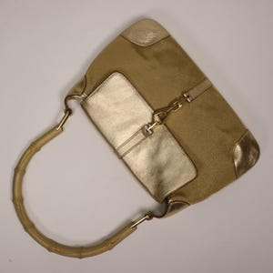 May include: A tan handbag with gold accents. The bag features a bamboo handle and gold-colored metal hardware. The front of the bag has a large gold-colored panel. The corners of the bag are also gold.