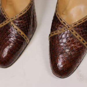K&ouml;nnte beinhalten: Ein Paar braune Lederschuhe mit Schlangenhautmuster und beigefarbener Naht. Die Schuhe haben eine spitze Zehenpartie und einen niedrigen Absatz.
