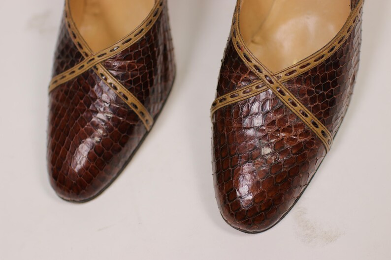 K&ouml;nnte beinhalten: Ein Paar braune Lederschuhe mit Schlangenhautmuster und beigefarbener Naht. Die Schuhe haben eine spitze Zehenpartie und einen niedrigen Absatz.