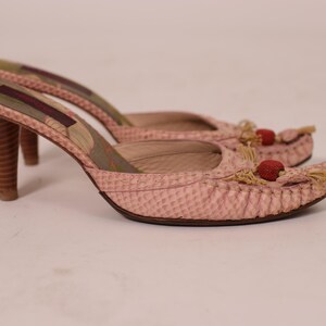 May include: A pair of pink high-heeled mules. The shoes have a textured pattern, wooden heels, and decorative tassels with red accents. The heels are approximately 3 inches tall.