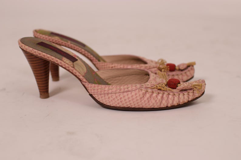May include: A pair of pink high-heeled mules. The shoes have a textured pattern, wooden heels, and decorative tassels with red accents. The heels are approximately 3 inches tall.