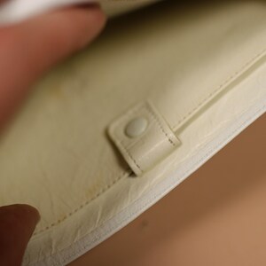 K&ouml;nnte beinhalten: Cremefarbenes Portemonnaie mit Druckknopfverschluss. Der Innenraum hat ein hellgelbes Futter und eine kleine Tasche. Das Portemonnaie ist aus einem glatten, m&ouml;glicherweise lederartigen Material gefertigt.
