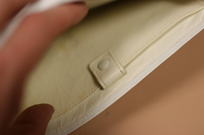 K&ouml;nnte beinhalten: Cremefarbenes Portemonnaie mit Druckknopfverschluss. Der Innenraum hat ein hellgelbes Futter und eine kleine Tasche. Das Portemonnaie ist aus einem glatten, m&ouml;glicherweise lederartigen Material gefertigt.