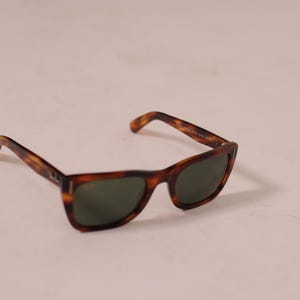 Puede incluir: Gafas de sol con un cl&aacute;sico estampado de carey. Las monturas son marrones y &aacute;mbar, con lentes de color verde oscuro. Las gafas de sol est&aacute;n ligeramente anguladas, mostrando la forma de las lentes y las patillas.