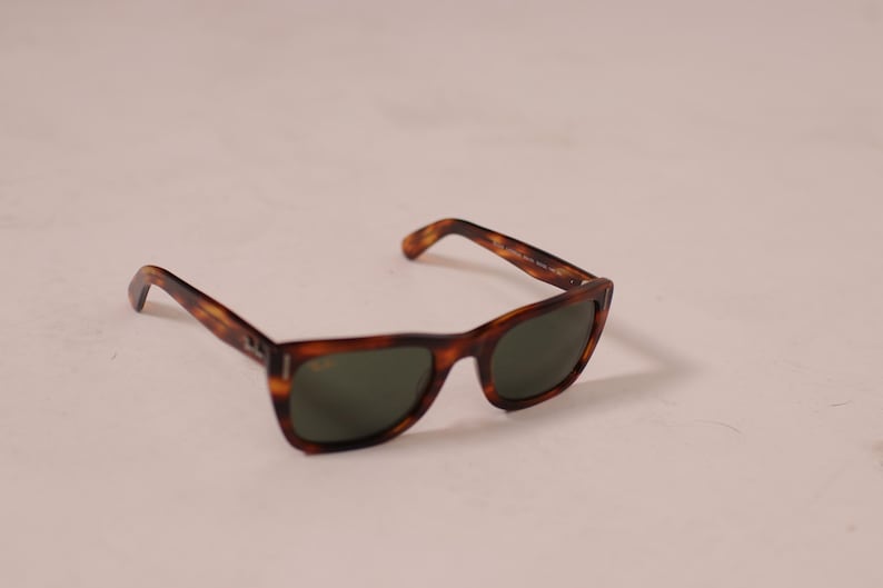 Puede incluir: Gafas de sol con un cl&aacute;sico estampado de carey. Las monturas son marrones y &aacute;mbar, con lentes de color verde oscuro. Las gafas de sol est&aacute;n ligeramente anguladas, mostrando la forma de las lentes y las patillas.