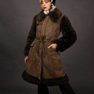 May include: A brown suede coat with a fur collar and cuffs. The coat has a belt and is lined with fur. The model is wearing a black and white fur hat.