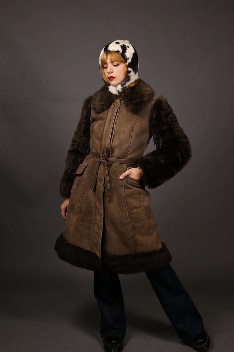 May include: A brown suede coat with a fur collar and cuffs. The coat has a belt and is lined with fur. The model is wearing a black and white fur hat.