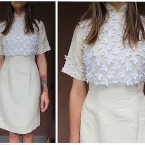 May include: A cream-colored dress with a white floral design on the bodice. The dress has a mandarin collar and short sleeves.