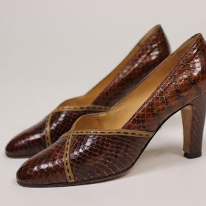 Può includere: Un paio di décolleté marroni in pelle di serpente con un bordo in pelle beige. Le scarpe hanno una punta a punta e un tacco alto.