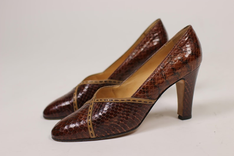 K&ouml;nnte beinhalten: Ein Paar braune Pumps aus Schlangenleder mit einem braunen Lederbesatz. Die Schuhe haben eine spitze Zehenpartie und einen hohen Absatz.