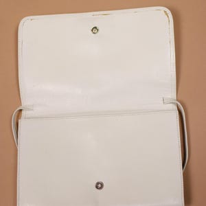 K&ouml;nnte beinhalten: Ge&ouml;ffnete, cremefarbene rechteckige Handtasche mit langem Riemen. Die Tasche hat einen Klappenverschluss mit einem Metallschnappverschluss. Das Innere ist sichtbar, und die Tasche scheint aus einem glatten Material zu bestehen.