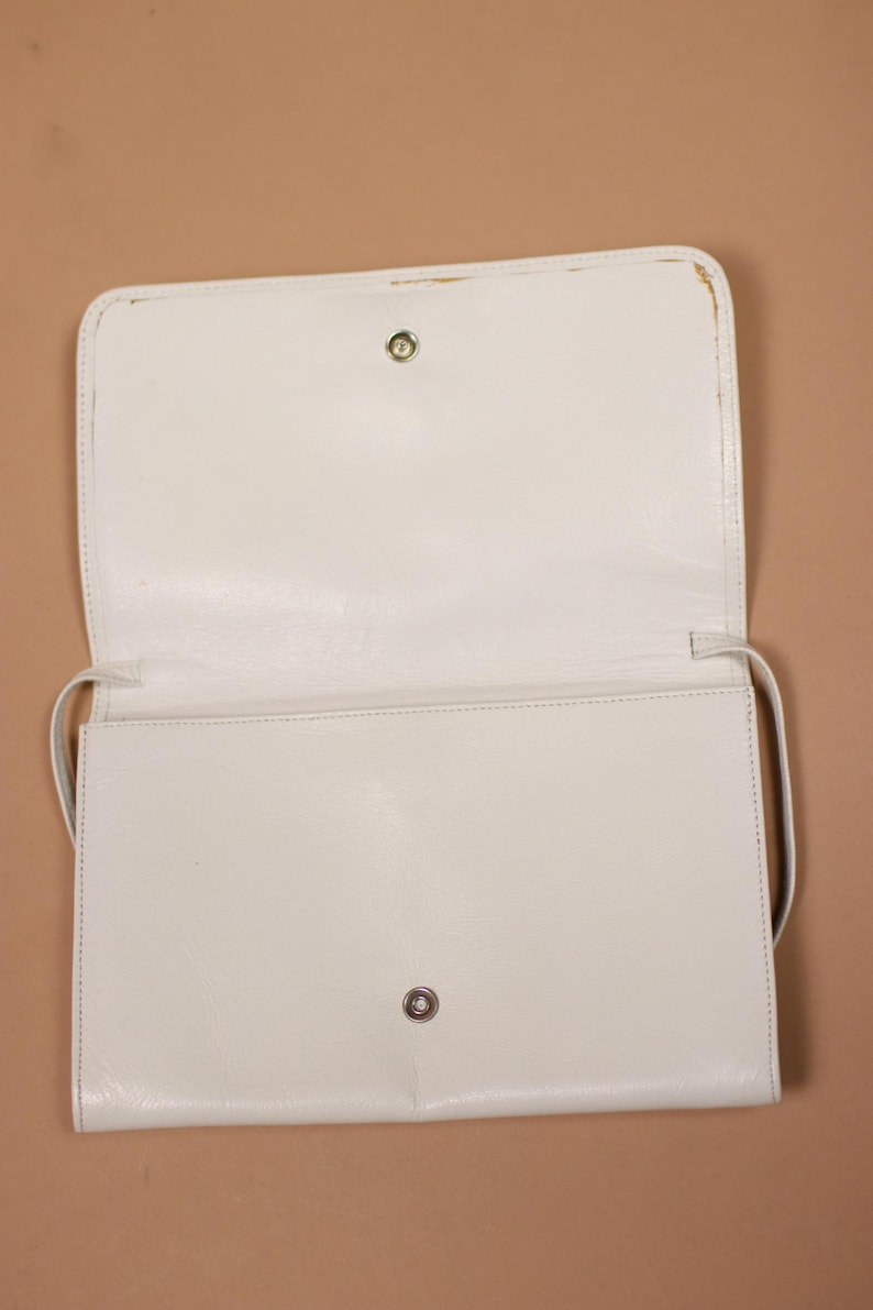K&ouml;nnte beinhalten: Ge&ouml;ffnete, cremefarbene rechteckige Handtasche mit langem Riemen. Die Tasche hat einen Klappenverschluss mit einem Metallschnappverschluss. Das Innere ist sichtbar, und die Tasche scheint aus einem glatten Material zu bestehen.