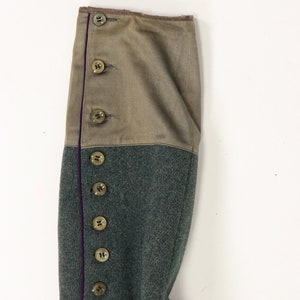 May include: Close-up of the sleeve of a green military jacket with a brown fabric cuff and a row of buttons down the side. The cuff has a purple trim.