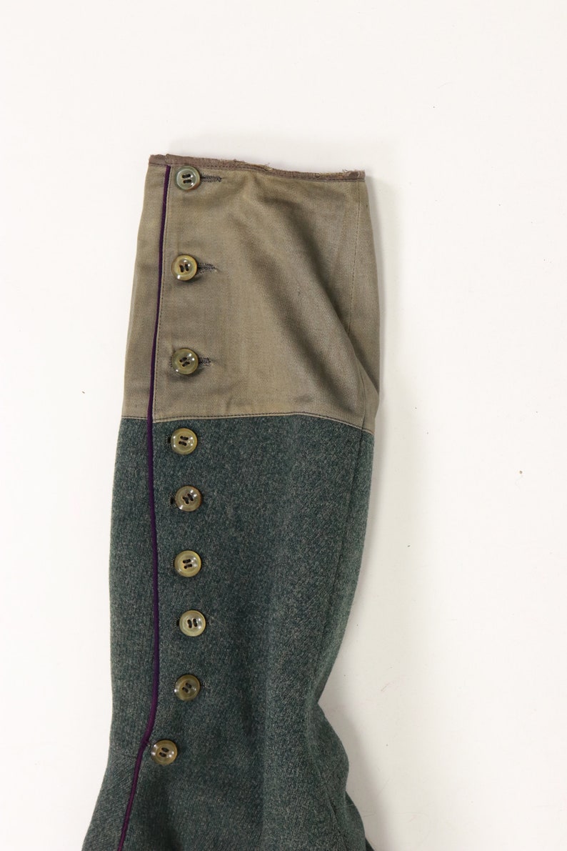 May include: Close-up of the sleeve of a green military jacket with a brown fabric cuff and a row of buttons down the side. The cuff has a purple trim.