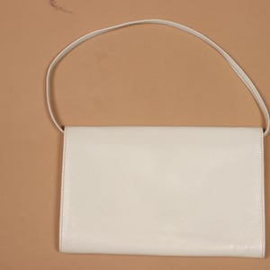 K&ouml;nnte beinhalten: Eine cremefarbene, rechteckige Handtasche mit einem d&uuml;nnen Schulterriemen. Die Tasche hat ein schlichtes, minimalistisches Design mit einem Klappenverschluss. Der Hintergrund ist einfarbig beige.