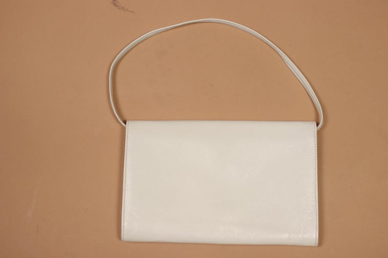 K&ouml;nnte beinhalten: Eine cremefarbene, rechteckige Handtasche mit einem d&uuml;nnen Schulterriemen. Die Tasche hat ein schlichtes, minimalistisches Design mit einem Klappenverschluss. Der Hintergrund ist einfarbig beige.
