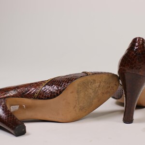 K&ouml;nnte beinhalten: Ein Paar braune Lederschuhe mit Absatz und Schlangenhautmuster. Die Schuhe haben eine spitze Zehenpartie und einen hohen Absatz.