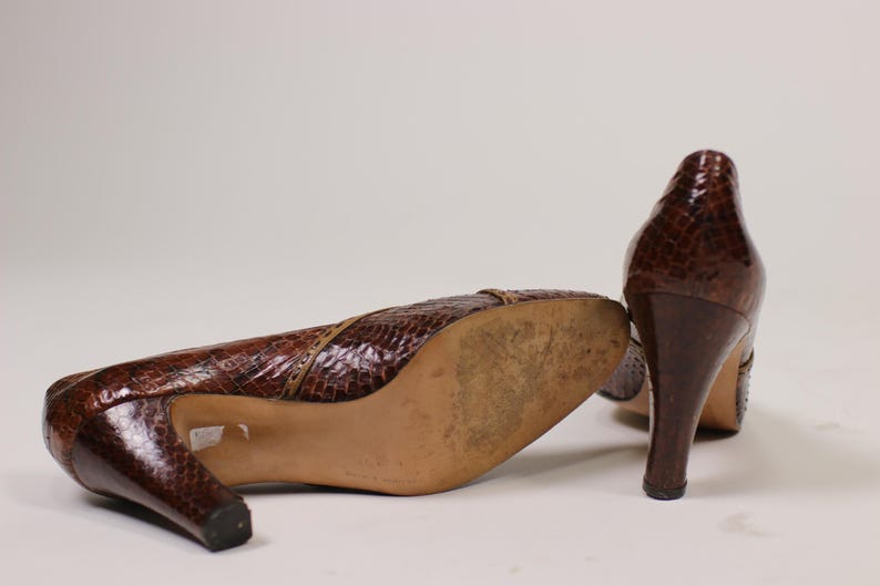 K&ouml;nnte beinhalten: Ein Paar braune Lederschuhe mit Absatz und Schlangenhautmuster. Die Schuhe haben eine spitze Zehenpartie und einen hohen Absatz.