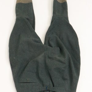 May include: A pair of dark green wool trousers with brown leather accents at the top of the legs. The trousers have a button closure at the waist and a pocket on the left side.