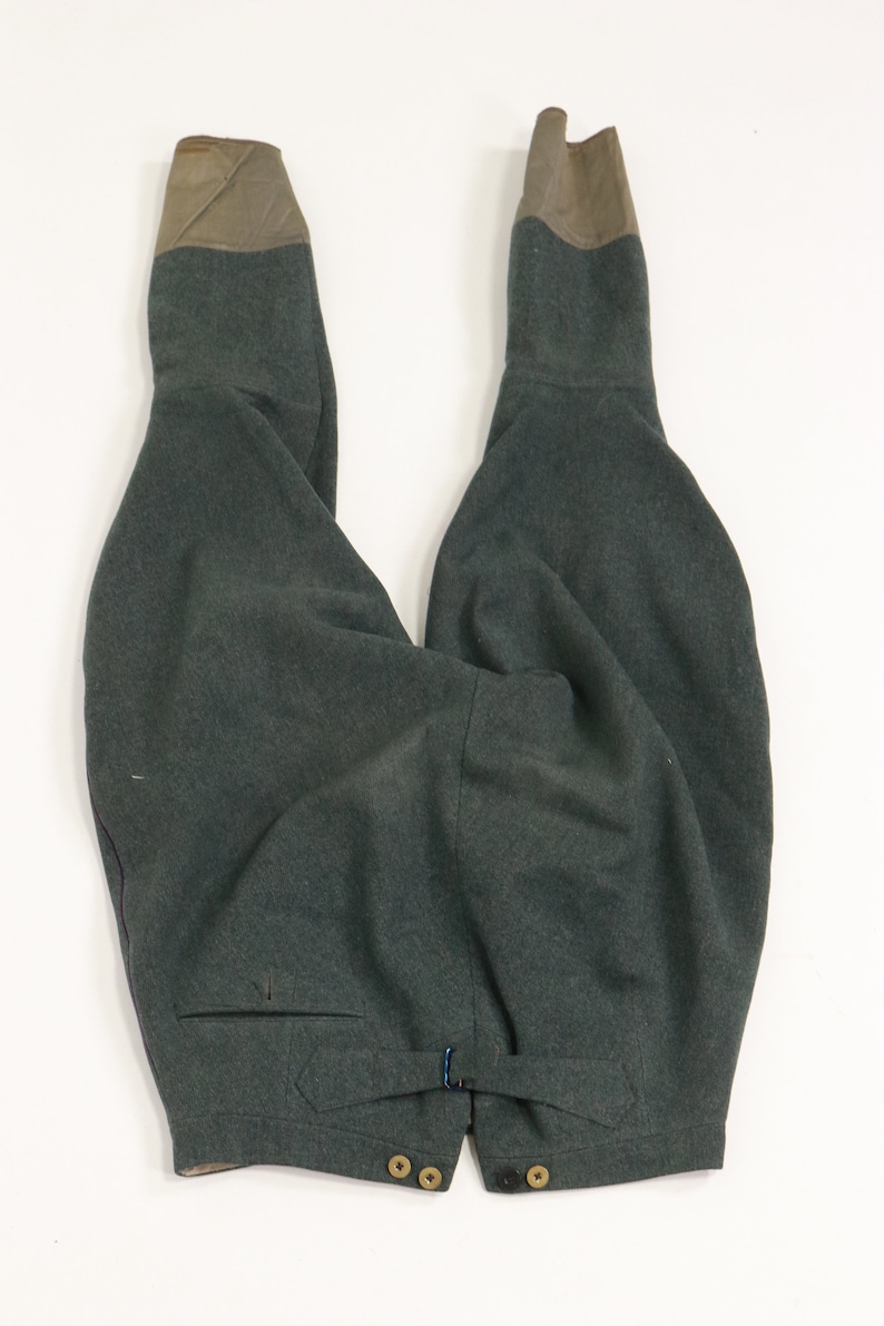 May include: A pair of dark green wool trousers with brown leather accents at the top of the legs. The trousers have a button closure at the waist and a pocket on the left side.