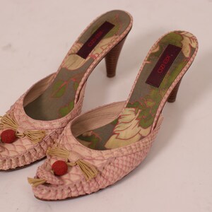 May include: A pair of pink, heeled mules with a textured, snakeskin-like pattern. The shoes feature small tassels, red accents, and floral patterned insoles. The heels are approximately 3 inches tall.