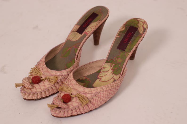 May include: A pair of pink, heeled mules with a textured, snakeskin-like pattern. The shoes feature small tassels, red accents, and floral patterned insoles. The heels are approximately 3 inches tall.