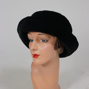 Puede incluir: Un sombrero cloche de terciopelo negro con un ala ancha y una corona redonda. El sombrero está sobre una cabeza de maniquí.