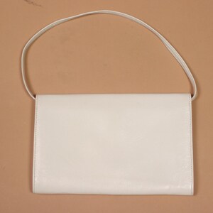 K&ouml;nnte beinhalten: Eine rechteckige, cremefarbene Handtasche mit einem d&uuml;nnen, passenden Riemen. Die Tasche hat ein schlichtes, klares Design mit einem Klappenverschluss. Der Hintergrund ist einfarbig hellbraun.
