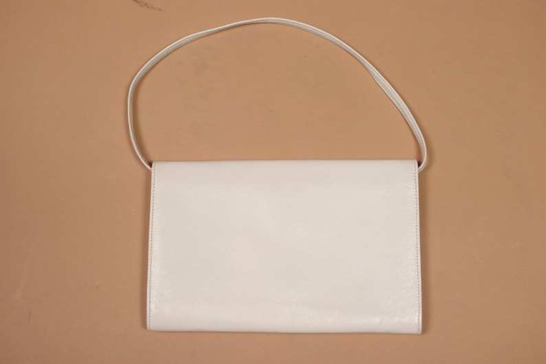 K&ouml;nnte beinhalten: Eine rechteckige, cremefarbene Handtasche mit einem d&uuml;nnen, passenden Riemen. Die Tasche hat ein schlichtes, klares Design mit einem Klappenverschluss. Der Hintergrund ist einfarbig hellbraun.