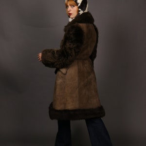 May include: A brown suede and shearling coat with a fur collar and cuffs. The coat has a belt and is worn over blue jeans.
