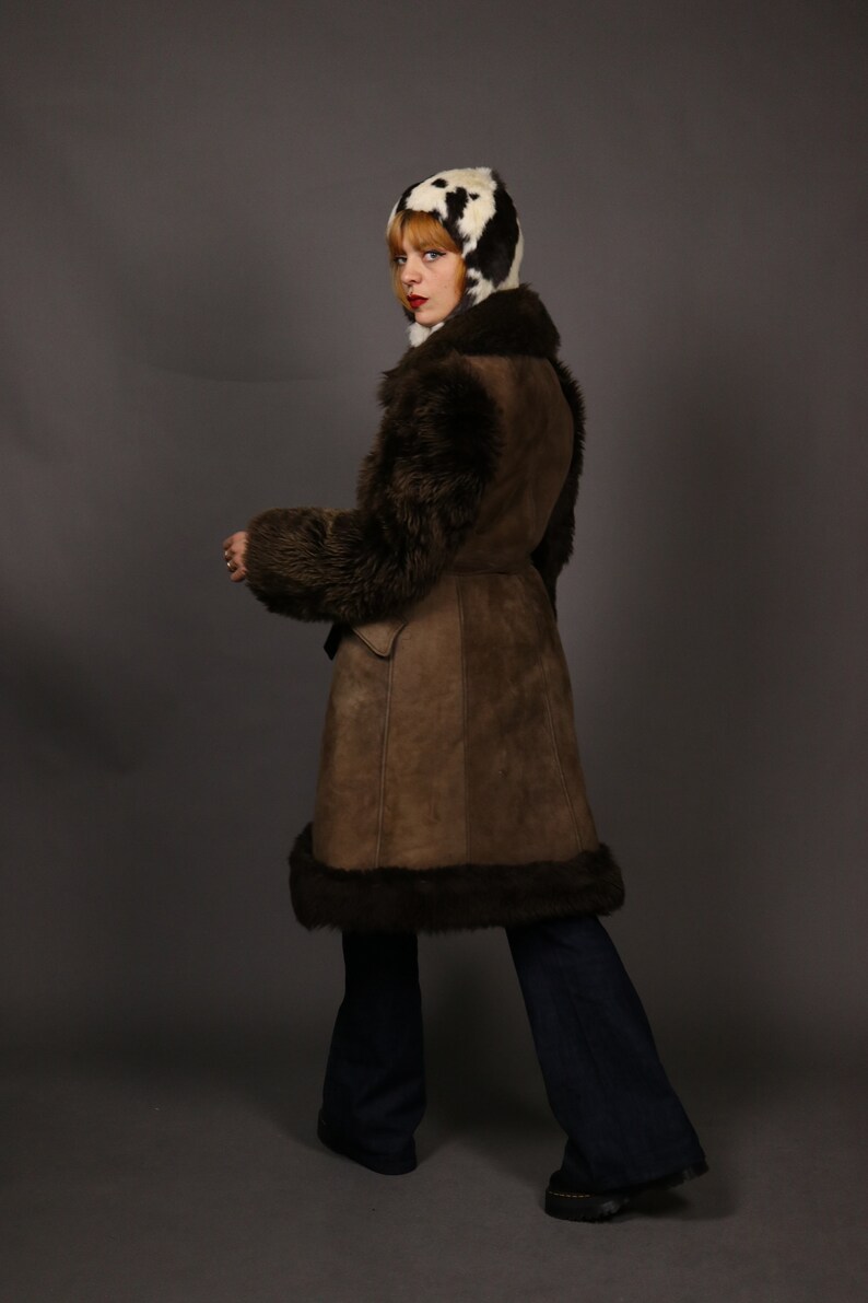 May include: A brown suede and shearling coat with a fur collar and cuffs. The coat has a belt and is worn over blue jeans.