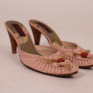 May include: A pair of pink heeled mules with a snakeskin pattern. The shoes have wooden heels, decorative tassels, and small red embellishments. The interior features a floral design.