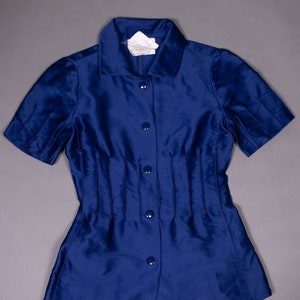 May include: A blue short-sleeved button-down shirt with a collar and a fitted waist. The shirt has a label that reads "Made in Italy, 100% Silk, Dry Clean Only".