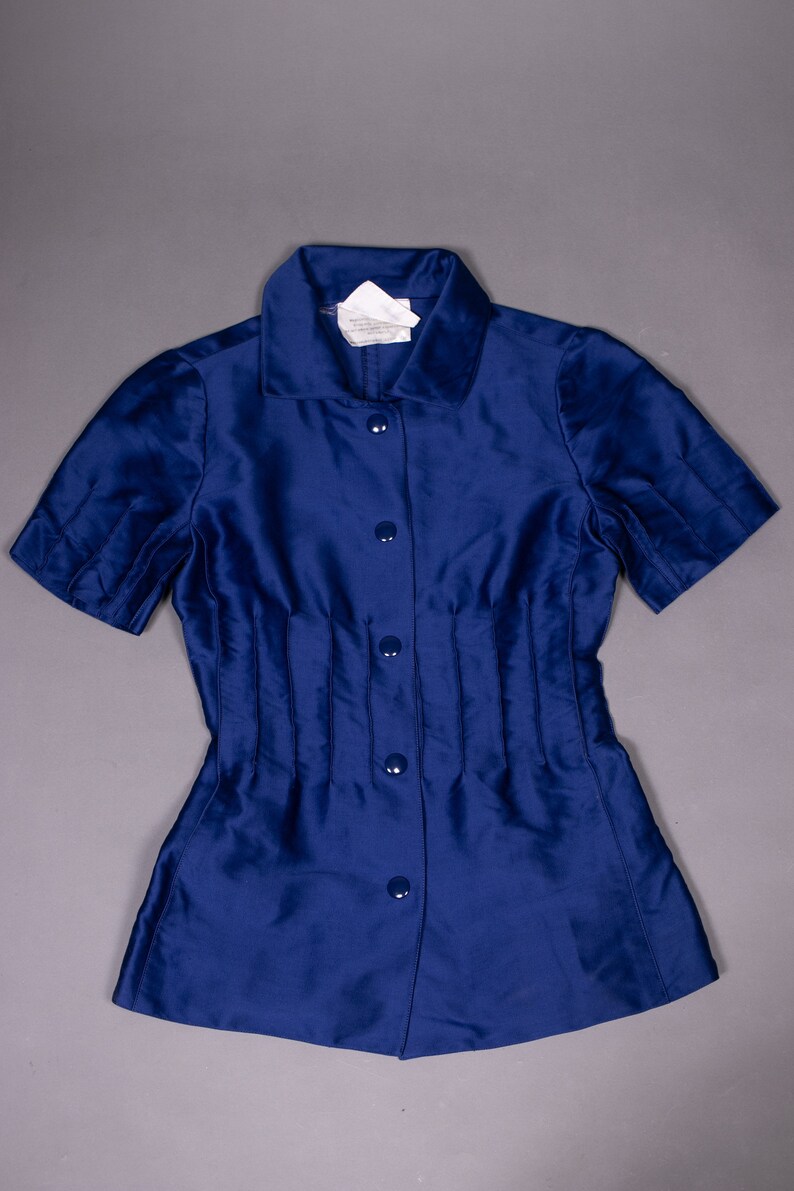 May include: A blue short-sleeved button-down shirt with a collar and a fitted waist. The shirt has a label that reads "Made in Italy, 100% Silk, Dry Clean Only".