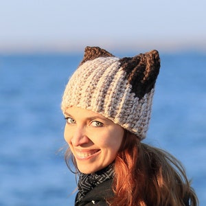 May include: A woman wearing a beige and brown knitted hat with cat ears. The hat is crocheted and has a textured pattern. The woman is smiling and looking to the side. She is standing in front of a blue body of water.