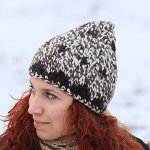 Puede incluir: Una mujer con el pelo largo y rizado rojo lleva un gorro de punto marrón y blanco y una bufanda de ganchillo. Está de pie en un entorno exterior nevado.