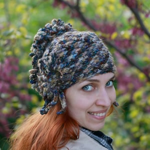 Puede incluir: Una mujer con el pelo largo y rojo lleva un gorro de punto marrón, azul y blanco con un diseño único. Lleva una chaqueta beige y está de pie frente a un fondo de flores rosas y verdes.