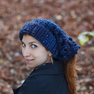 Puede incluir: Una mujer con el pelo largo y rojo lleva una boina de punto azul. Lleva una chaqueta negra y mira por encima del hombro.