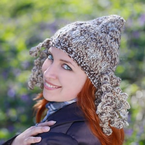 May include: A woman with long red hair wearing a brown and white crocheted hat with a unique design. She is wearing a dark blue jacket and has a scarf around her neck.