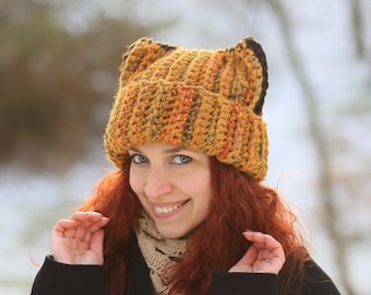 May include: A woman with long red hair is wearing a crocheted hat with cat ears. The hat is orange and brown with a striped pattern. She is wearing a black coat and a cream-colored scarf.