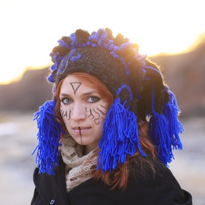 Puede incluir: Una mujer con un sombrero negro de ganchillo con borlas de hilo azul y una chaqueta negra. Tiene un diseño geométrico dibujado en la frente y sombra de ojos azul.