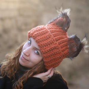 May include: An orange crocheted hat with two faux fur accents. The hat is a warm, earthy tone, ideal for cold weather. The hat is made of a thick yarn, and the faux fur adds a touch of luxury. The hat is a stylish accessory for any outfit.