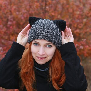 May include: A woman with long red hair wearing a black sweater and a gray and black knit hat with cat ears.