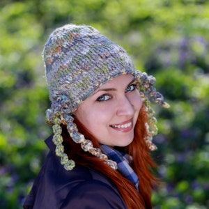 Puede incluir: Una mujer con cabello rojo lleva un gorro de punto gris, verde y marrón con largas orejeras de ganchillo. Está sonriendo y mirando a la cámara.