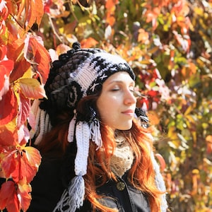 Puede incluir: Una mujer con el pelo largo y rojo lleva un gorro de ganchillo blanco y negro con una larga franja. Está de pie frente a una pared de hojas rojas y amarillas.
